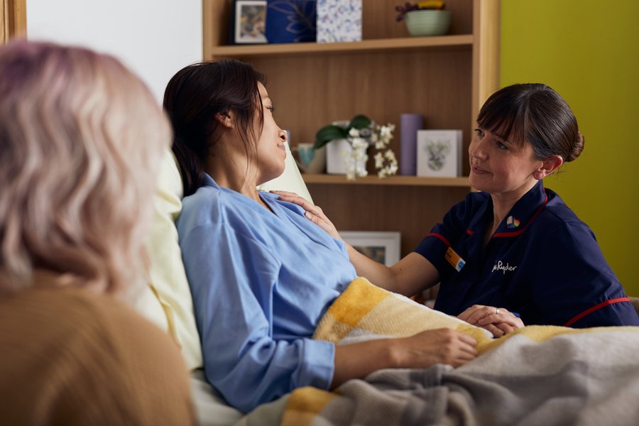 A nurse in a dark blue uniform is sat next to a bed, where a female patient is sitting. In the foreground is the back of another woman.