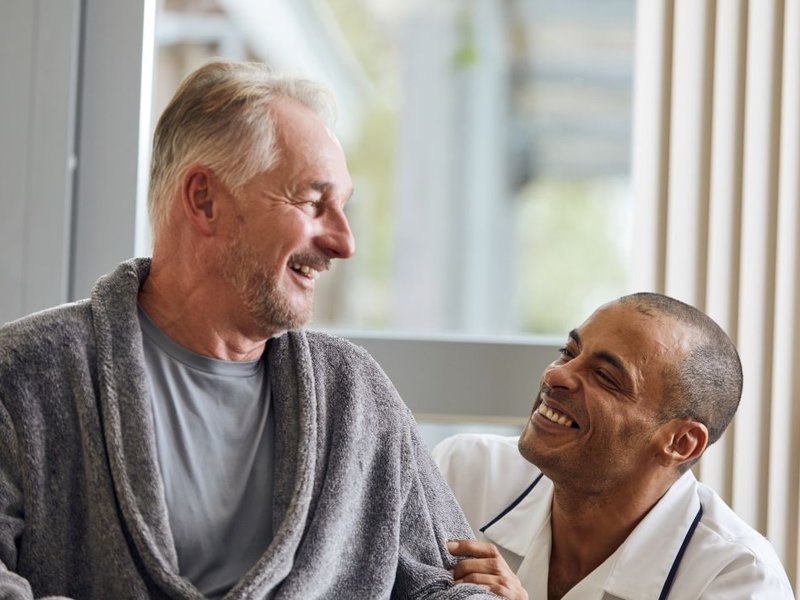 Man sitting in grey dressing gown smiles down at man in white nurses uniform.